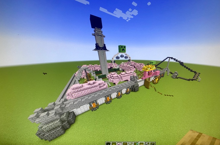 Na zdjęciu widzimy park rozrywki zbudowany w grze Minecraft. Konstrukcja zawiera:  Wieżę przypominającą wieżę widokową lub atrakcję typu "drop tower".  Rollercoaster po prawej stronie, z torami prowadzącymi w górę i w dół.  Motyw różowy, prawdopodobnie inspirowany tematyką słodyczy lub bajek – dużo różowych bloków, a na froncie znajduje się coś, co przypomina twarz postaci (może świnka lub inny kreskówkowy motyw).  Zielone twarze Creeperów, co sugeruje motyw Minecraftowy.  Łuk z napisem lub dekoracją na środku w tle, z Creeperem na górze