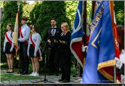 Na zdjęciu widzimy oficjalną uroczystość, prawdopodobnie związaną z obchodami patriotycznymi lub szkolnymi.  Oto szczegóły, które można zauważyć:  Po lewej stronie stoi poczet sztandarowy – młodzież ubrana galowo (białe koszule, ciemne spódnice/spodnie i czerwone szarfy) trzyma sztandar.  Na środku dwoje dorosłych (mężczyzna i kobieta) występuje przy mikrofonie, prawdopodobnie odczytując przemówienie lub fragmenty upamiętniające.