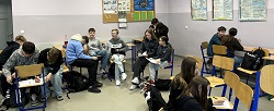 Na zdjęciu widać uczniów siedzących w grupach w sali lekcyjnej. Uczestniczą oni prawdopodobnie w warsztatach – rozmawiają ze sobą i robią notatki na kartkach. Atmosfera wygląda na swobodną i sprzyjającą współpracy. Można zauważyć plakaty edukacyjne na ścianach oraz typowe wyposażenie klasy: krzesła, ławki i tablicę.