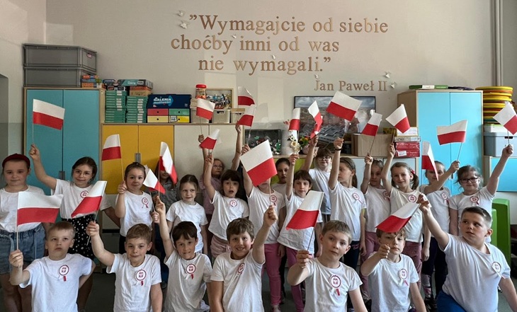 Na zdjęciu widać grupę dzieci w sali lekcyjnej, ubrane w białe koszulki z przypiętymi kokardkami narodowymi. Każde z dzieci trzyma w ręce małą polską flagę. W tle, na ścianie, widnieje cytat Jana Pawła II:  „Wymagajcie od siebie, choćby inni od was nie wymagali.” — Jan Paweł II