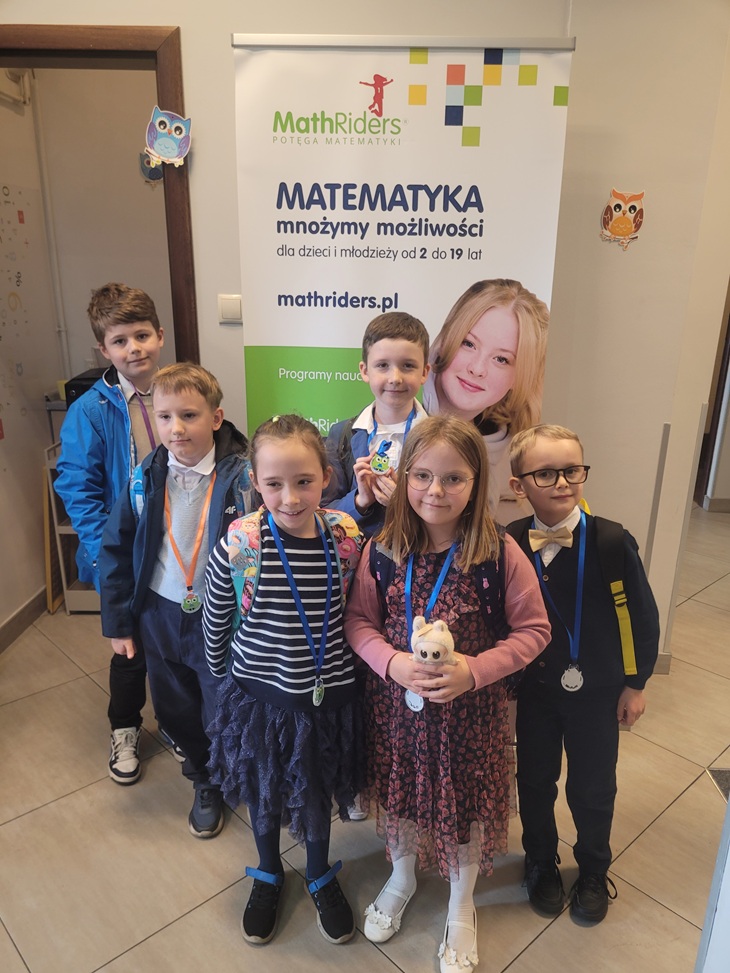 Na zdjęciu znajduje się sześciu młodych uczniów (prawdopodobnie z klas 1–3 szkoły podstawowej), którzy stoją przed roll-upem promującym program edukacyjny MathRiders. Wszystkie dzieci mają na szyjach medale, co sugeruje, że brały udział w konkursie matematycznym – najpewniej II etapie Ogólnopolskiego Konkursu Matematycznego "Puchacz Piotr".