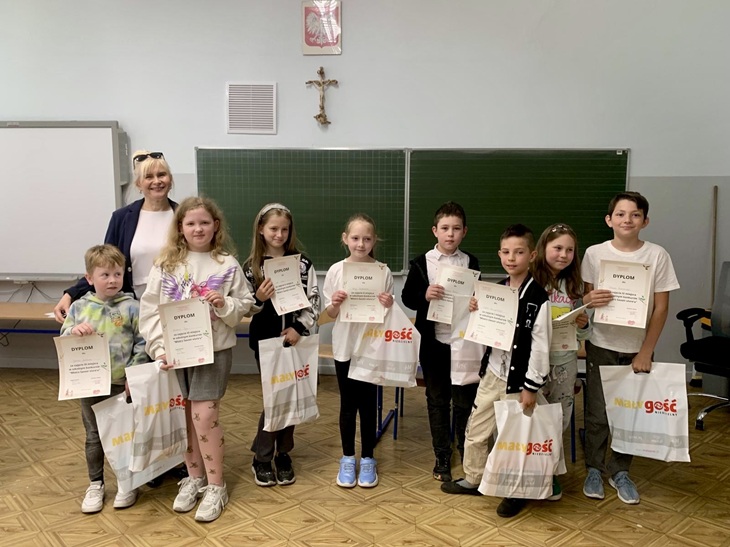 Na zdjęciu widzimy uczestników konkursu „Mistrz savoir-vivre’u”, o którym mowa w poprzednim tekście.  Uczniowie klas trzecich stoją w sali lekcyjnej, dumnie prezentując swoje dyplomy oraz nagrody za udział w konkursie. Towarzyszy im nauczycielka – najprawdopodobniej organizatorka wydarzenia. Dzieci ubrane są swobodnie, a na ich twarzach widać radość i satysfakcję z udziału w wydarzeniu. 