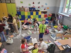 Na zdjęciu widać grupę dzieci w klasie szkolnej, które biorą udział w zajęciach związanych z recyklingiem lub Dniem Ziemi 🌍. Dzieci siedzą i pracują w grupach – wokół nich widać wiele materiałów wtórnych: papier, plastik, kartony, reklamówki itp. Część uczniów prawdopodobnie przygotowuje stroje lub prace plastyczne z surowców wtórnych, co sugeruje, że to może być pokaz eko-mody lub warsztaty ekologiczne.