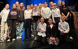 Na zdjęciu widzimy grupę uczniów – uczestników przeglądu teatralnego Perypetie 2025. Uczniowie stoją na scenie, uśmiechają się i trzymają dyplom oraz statuetkę, co świadczy o zdobyciu nagrody. W tle widać niebieską kurtynę teatralną, a uczniom towarzyszą nauczyciele lub opiekunowie. Grupa wygląda na zgraną i dumna ze swojego osiągnięcia – to z pewnością chwila radości po udanym występie!