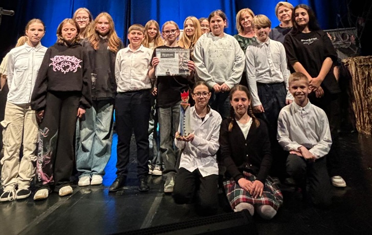 Na zdjęciu widzimy grupę uczniów – uczestników przeglądu teatralnego Perypetie 2025. Uczniowie stoją na scenie, uśmiechają się i trzymają dyplom oraz statuetkę, co świadczy o zdobyciu nagrody. W tle widać niebieską kurtynę teatralną, a uczniom towarzyszą nauczyciele lub opiekunowie. Grupa wygląda na zgraną i dumna ze swojego osiągnięcia – to z pewnością chwila radości po udanym występie!