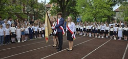 Na zdjęciu widzimy uroczystość szkolną, najprawdopodobniej zakończenie roku szkolnego. Uczniowie ubrani są odświętnie – chłopcy w białych koszulach, dziewczynki w białych bluzkach i ciemnych spódniczkach.  Na pierwszym planie stoi poczet sztandarowy – trójka starszych uczniów (dwie dziewczyny i chłopiec), trzymających szkolny sztandar. W tle widać grupy uczniów i nauczycieli ustawionych w szeregu na boisku szkolnym, prawdopodobnie podczas hymnu lub innego oficjalnego momentu uroczystości.