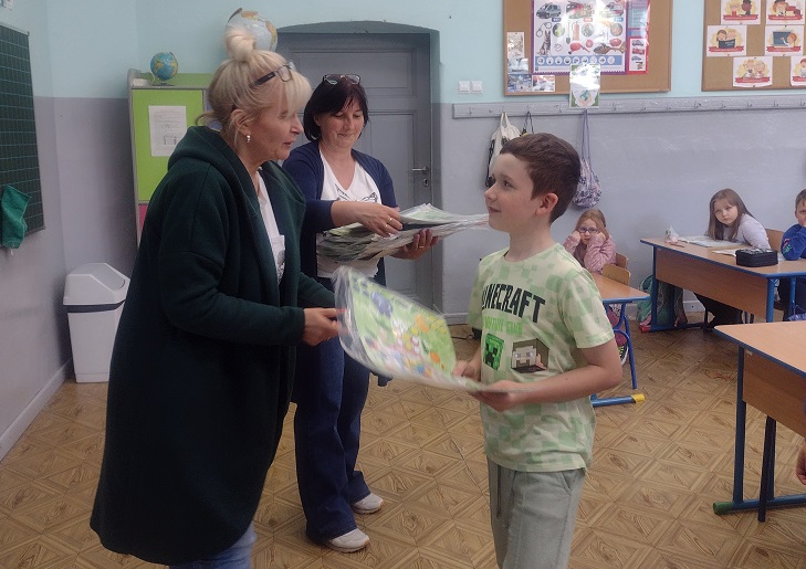 Na zdjęciu widać moment wręczania nagród uczniowi przez nauczycielkę podczas szkolnego konkursu. W tle znajdują się inni uczniowie siedzący w ławkach oraz druga nauczycielka trzymająca prace konkursowe.