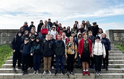 Na zdjęciu widać dużą grupę uczniów i kilku dorosłych opiekunów ustawionych na szerokich, kamiennych schodach w plenerze. Za nimi rozciąga się niskie kamienne ogrodzenie i niebo z lekkim zachmurzeniem. Większość osób ma na sobie kurtki i bluzy, co sugeruje chłodniejszą pogodę. Grupa pozuje do wspólnej fotografii, uśmiechając się i patrząc w stronę aparatu, tworząc pamiątkowe ujęcie z wycieczki.