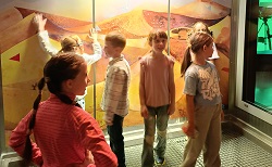 Na zdjęciu widać grupę dzieci znajdujących się w pomieszczeniu przypominającym interaktywne muzeum lub planetarium. Dzieci stoją na metalowej kratownicy, a za nimi znajduje się kolorowa ściana przedstawiająca fantazyjny, kosmiczny lub pustynny krajobraz — z wzgórzami w odcieniach pomarańczu, żółci i fioletu oraz abstrakcyjnymi elementami graficznymi (np. czerwone trójkąty).