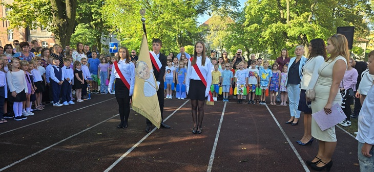 Na zdjęciu widzimy uroczystość rozpoczęcia roku szkolnego, która odbywa się na boisku szkolnym.  Na pierwszym planie stoi poczet sztandarowy – troje uczniów w galowych strojach, z biało-czerwonymi szarfami. Jeden z nich trzyma sztandar szkoły. Obok znajdują się nauczyciele oraz Pani Dyrektor prowadząca uroczystość.  W tle zgromadzeni są uczniowie