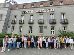 Na zdjęciu widać grupę dzieci stojących przed budynkiem z dużym napisem „TEATR MIEJSKI”. Uczniowie ustawieni są w rzędzie, uśmiechają się i pozują do wspólnej fotografii. Za nimi widać szare elewacje teatru z kilkoma wysokimi oknami i małymi balkonami. Wokół budynku rosną krzewy, a na pierwszym planie widać chodnik z kostki brukowej. Zdjęcie sprawia wrażenie pamiątkowego ujęcia z wycieczki do teatru w chłodny, pochmurny dzień.