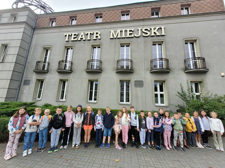 Na zdjęciu widać grupę dzieci stojących przed budynkiem z dużym napisem „TEATR MIEJSKI”. Uczniowie ustawieni są w rzędzie, uśmiechają się i pozują do wspólnej fotografii. Za nimi widać szare elewacje teatru z kilkoma wysokimi oknami i małymi balkonami. Wokół budynku rosną krzewy, a na pierwszym planie widać chodnik z kostki brukowej. Zdjęcie sprawia wrażenie pamiątkowego ujęcia z wycieczki do teatru w chłodny, pochmurny dzień.