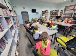 uczniowie w bibliotece siedzą za stołami, słuchają nauczycielki