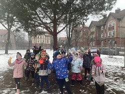 Na zdjęciu widać grupę dzieci ubranych w zimowe kurtki i czapki, stojących na ośnieżonym terenie z drzewami. Dzieci trzymają w rękach jedzenie dla ptaków i wyglądają na radosne, jak podczas wspólnej akcji na świeżym powietrzu.