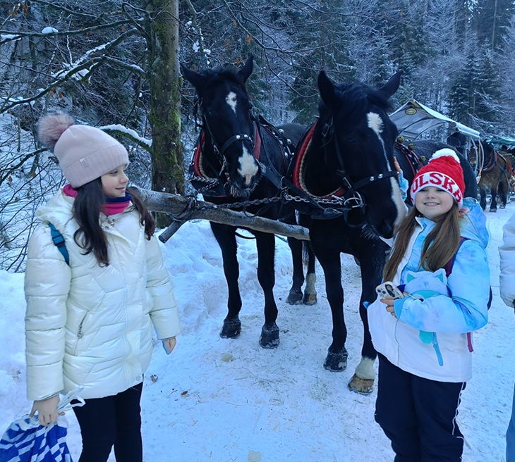 Na zdjęciu widać dwoje dzieci stojących na ośnieżonej leśnej drodze obok dwóch zaprzęgniętych koni. Wokół jest zimowy krajobraz – drzewa pokryte śniegiem i sanie w tle. Dzieci są ciepło ubrane w zimowe kurtki i czapki, a cała scena ma klimat radosnej, zimowej wycieczki.