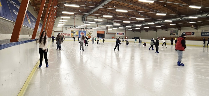 Na zdjęciu widać dużą halę lodowiska, na którego tafli jednocześnie jeździ wiele osób. Uczniowie poruszają się po lodzie w różnych kierunkach, część z nich jeździ samodzielnie, inni w mniejszych grupach. Widoczna jest aktywność i ruch, a cała scena oddaje atmosferę wspólnej zabawy oraz sportowego wysiłku.  Lodowisko jest przestronne i dobrze oświetlone, a bandy oraz konstrukcja hali wskazują na obiekt miejski przystosowany do zajęć rekreacyjnych. Zdjęcie pokazuje radosny, aktywny charakter wyjścia szkolnego i czas spędzony na świeżej aktywności.