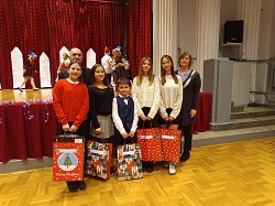 Uczniowie z nauczycielka stoja na tle sceny, w rękach trzymaja torby z upominkami, są odświętnie ubrani