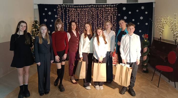 Na zdjęciu widać grupę uczniów i nauczycieli pozujących wspólnie na tle świątecznej dekoracji 🎄✨ Uczestnicy są elegancko ubrani, a część z nich trzyma papierowe torby z nagrodami 🎁📸 Atmosfera jest uroczysta i radosna.