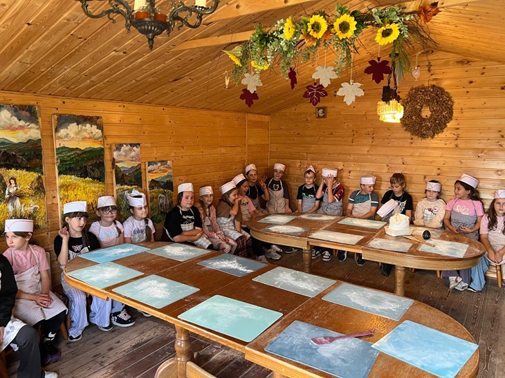 Na zdjęciu widać grupę dzieci uczestniczących w zajęciach edukacyjnych w drewnianej, przytulnej chacie. Dzieci siedzą przy stołach, na których przygotowano materiały potrzebne do zajęć. Uczestnicy mają na głowach charakterystyczne papierowe czapeczki, co podkreśla warsztatowy charakter spotkania. Wnętrze chaty jest ozdobione jesiennymi dekoracjami, m.in. słonecznikami, liśćmi i innymi naturalnymi elementami. Na ścianach znajdują się obrazy przedstawiające wiejskie i przyrodnicze krajobrazy.
