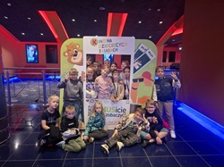 Na zdjęciu widać grupę dzieci pozujących do wspólnej fotografii w kolorowym, nowoczesnym wnętrzu, prawdopodobnie w centrum rozrywki. Dzieci siedzą i stoją wokół dużej, ozdobnej planszy fotograficznej z napisem „Kino na dziecięcych zasadach”.  W tle znajduje się ciemne pomieszczenie z efektownym oświetleniem — dominują czerwone i niebieskie światła oraz dekoracje nawiązujące do kina. Dzieci są uśmiechnięte i wyglądają na zadowolone ze wspólnego wyjścia. Całość sprawia wrażenie pamiątkowego zdjęcia z grupowej wycieczki lub wydarzenia dla dzieci.