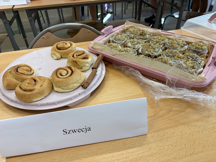Na zdjęciu widać stół przygotowany na szkolną degustację z okazji Europejskiego Dnia Języków. Na białym talerzu znajdują się szwedzkie cynamonki, a obok duża blacha z innym wypiekiem posypanym kruszonką. Przed potrawami ustawiono kartkę z napisem „Szwecja”, wskazującą kraj, którego kuchnię reprezentują przygotowane smakołyki. W tle widać szkolne ławki i krzesła.