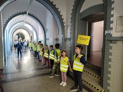 Na zdjęciu widać grupę uczniów ustawionych w szeregu na szkolnym korytarzu. Dzieci mają na sobie jaskrawe kamizelki odblaskowe, nawiązujące do działań związanych z bezpieczeństwem na drodze.  Najmłodsi uczniowie trzymają się za ręce i tworzą „odblaskowy łańcuch”, który ciągnie się wzdłuż korytarza. Jeden z chłopców trzyma żółtą tabliczkę z hasłem „Świeć przykładem na drodze!”. W tle widać kolejnych uczniów uczestniczących w akcji.  Zdjęcie pokazuje zaangażowanie dzieci w szkolną inicjatywę promującą widoczność i bezpieczne zachowanie na drodze.