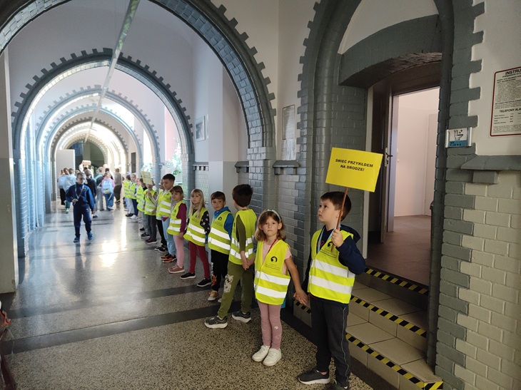 Na zdjęciu widać grupę uczniów ustawionych w szeregu na szkolnym korytarzu. Dzieci mają na sobie jaskrawe kamizelki odblaskowe, nawiązujące do działań związanych z bezpieczeństwem na drodze.  Najmłodsi uczniowie trzymają się za ręce i tworzą „odblaskowy łańcuch”, który ciągnie się wzdłuż korytarza. Jeden z chłopców trzyma żółtą tabliczkę z hasłem „Świeć przykładem na drodze!”. W tle widać kolejnych uczniów uczestniczących w akcji.  Zdjęcie pokazuje zaangażowanie dzieci w szkolną inicjatywę promującą widoczność i bezpieczne zachowanie na drodze.
