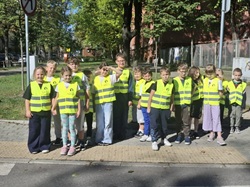 Na zdjęciu widać grupę uczniów stojących na chodniku przy ulicy. Dzieci mają na sobie jaskrawożółte kamizelki odblaskowe, dzięki którym są dobrze widoczne dla kierowców.