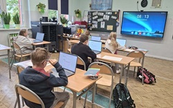 Na zdjęciu widać salę lekcyjną, w której uczniowie siedzą w ławkach i pracują przy laptopach. Każdy z nich skupia się na wykonywaniu zadań na komputerze. W klasie znajdują się drewniane ławki, krzesła, szafki oraz tablica z materiałami edukacyjnymi. Na ścianie wisi duży ekran multimedialny, który wyświetla godzinę. Duże okna zapewniają dostęp do naturalnego światła, a rośliny doniczkowe i dekoracje tworzą przyjemną atmosferę. Widać również plecaki i przybory szkolne. Zdjęcie przedstawia nowoczesną lekcję, podczas której uczniowie wykorzystują technologię do nauki.