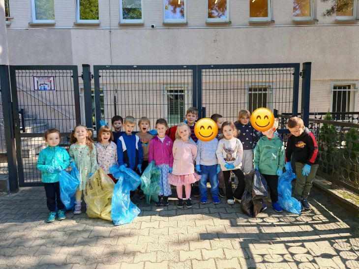 grupa dzieci stojąca przed przedszkolem z workami w rękach, gotowe do sprzątania