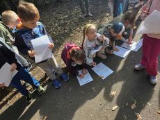 grupa dzieci na dworze, kucająca i rozwiązująca zadania na kartce
