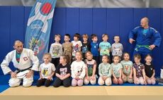 grupa dzieci ubrana w sportrowe stroje stojąca na sali gimnastycznej z trenerami judo ubranymi w szaty do judo.