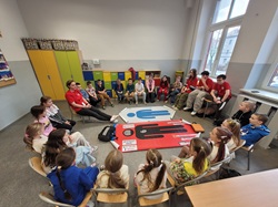 Uczniowie klasy 2b siedzą w kręgu podczas zajęć z pierwszej pomocy. Na środku znajduje się mata szkoleniowa do nauki RKO, a prowadzący omawia zasady udzielania pomocy. Dzieci aktywnie słuchają i uczestniczą w ćwiczeniach praktycznych.