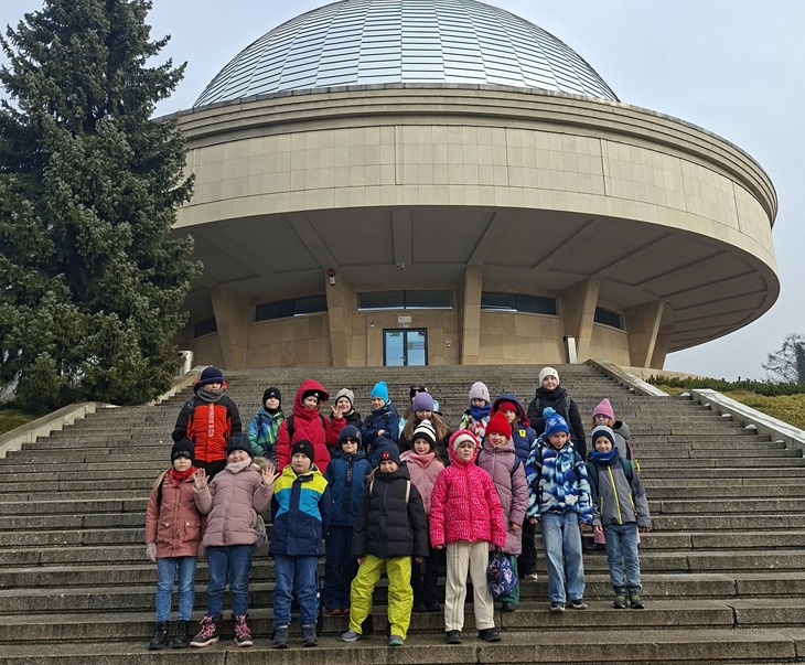 Na zdjęciu widać uczniów klas 2a i 2b stojących na schodach przed budynkiem planetarium z charakterystyczną kopułą. Dzieci są ciepło ubrane i pozują do wspólnej fotografii, która dokumentuje ich wizytę oraz udział w edukacyjnej wycieczce.