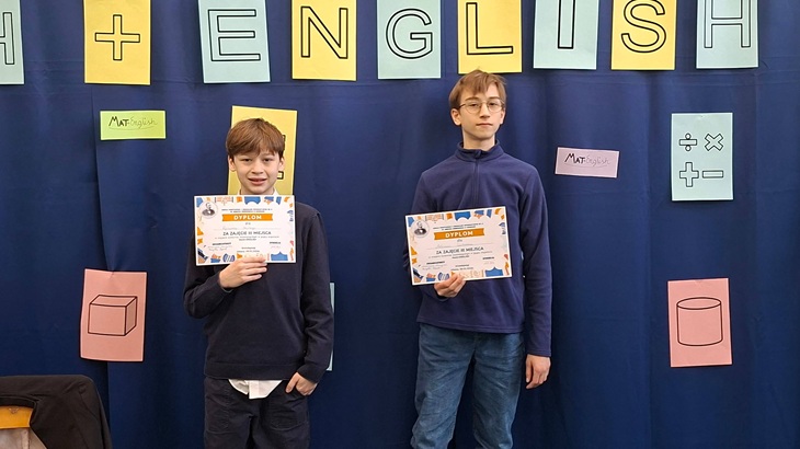 Na zdjęciu widać dwóch uczniów stojących na tle szkolnej gazetki tematycznej związanej z matematyką i językiem angielskim („Math + English”). Chłopcy trzymają dyplomy potwierdzające zdobycie III miejsca w konkursie matematycznym w języku angielskim. W tle znajdują się kolorowe plansze z symbolami matematycznymi i bryłami, podkreślające edukacyjny charakter wydarzenia.
