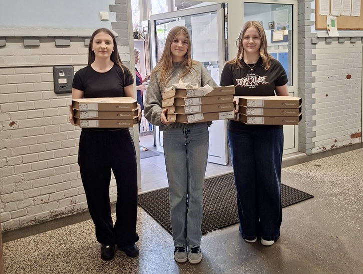 Na zdjęciu stoją trzy młode osoby w korytarzu (prawdopodobnie w szkole lub budynku publicznym). Każda z nich trzyma w rękach kilka pudełek z pizzą ułożonych jedno na drugim. Stoją obok siebie i patrzą w stronę aparatu. W tle widać jasne ściany z cegły, drzwi oraz tablicę z ogłoszeniami.