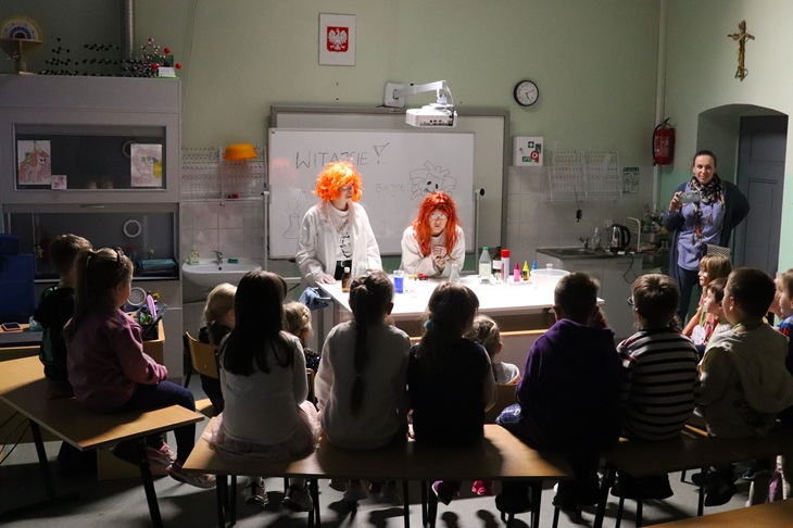 Na zdjęciu widać zajęcia dla dzieci podczas szkolnego wydarzenia. Dwóch „szalonych naukowców” w kolorowych perukach prowadzi eksperymenty przy stole, a uczniowie siedzą naprzeciwko i z zaciekawieniem obserwują pokaz. W tle znajduje się sala lekcyjna z tablicą i pomocami naukowymi, a całość ma radosną i edukacyjną atmosferę.