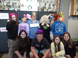 Na zdjęciu widać grupę uczniów wraz z nauczycielką, którzy pozują do zdjęcia podczas szkolnych obchodów Międzynarodowego Dnia Języka Ojczystego. Uczestnicy mają na sobie różne zabawne rekwizyty, kapelusze i maski, co nadaje wydarzeniu radosny i kreatywny charakter. Fotografia została wykonana przy specjalnej „ściance”, gdzie uczniowie mogli poczuć się wyjątkowo i zrobić pamiątkowe zdjęcie.