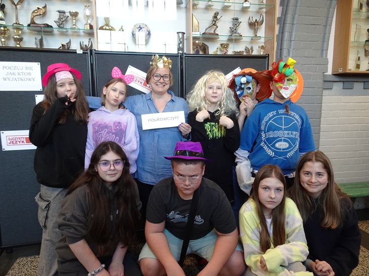 Na zdjęciu widać grupę uczniów wraz z nauczycielką, którzy pozują do zdjęcia podczas szkolnych obchodów Międzynarodowego Dnia Języka Ojczystego. Uczestnicy mają na sobie różne zabawne rekwizyty, kapelusze i maski, co nadaje wydarzeniu radosny i kreatywny charakter. Fotografia została wykonana przy specjalnej „ściance”, gdzie uczniowie mogli poczuć się wyjątkowo i zrobić pamiątkowe zdjęcie.