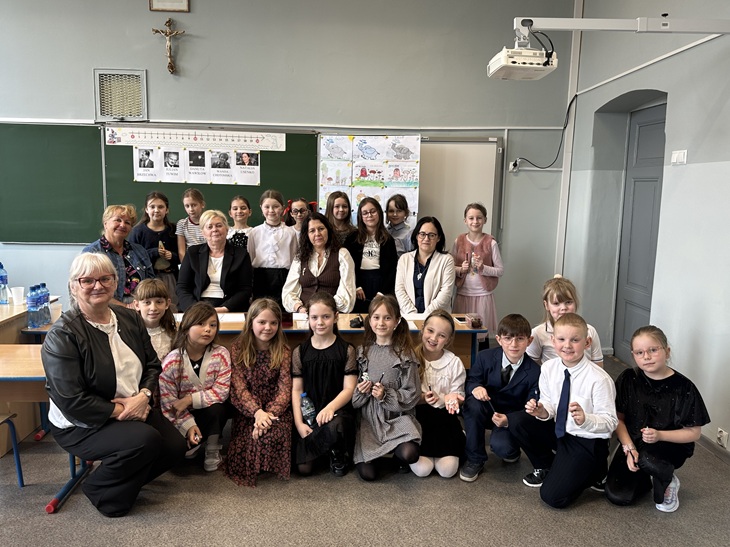 Na zdjęciu widać grupę uczniów oraz nauczycieli w sali lekcyjnej. Wszyscy pozują do wspólnego zdjęcia po wydarzeniu szkolnym. Dzieci stoją i siedzą z przodu, a nauczyciele znajdują się za nimi. W tle widać tablicę szkolną z materiałami edukacyjnymi oraz projektor. Wszyscy są uśmiechnięci.