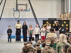 Na zdjęciu widać uczestników szkolnego konkursu „Mam Talent”, stojących na sali gimnastycznej przed publicznością. Na scenie ustawione są dzieci i młodzież, a w tle znajduje się dekoracja z gwiazdami. Widzowie siedzą naprzeciwko, uważnie obserwując występ, co tworzy atmosferę wspólnego świętowania i wsparcia