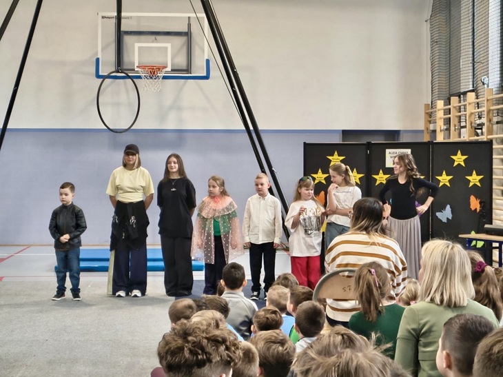 Na zdjęciu widać uczestników szkolnego konkursu „Mam Talent”, stojących na sali gimnastycznej przed publicznością. Na scenie ustawione są dzieci i młodzież, a w tle znajduje się dekoracja z gwiazdami. Widzowie siedzą naprzeciwko, uważnie obserwując występ, co tworzy atmosferę wspólnego świętowania i wsparcia