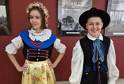Na zdjęciu widać dwoje uczniów prezentujących tradycyjne stroje śląskie. Dziewczynka ma na sobie kolorowy strój ludowy z białą bluzką, ozdobnym gorsetem, koralami oraz kwiatowym wiankiem na głowie. Jej strój uzupełnia jasna spódnica z kwiatowym wzorem. Chłopiec jest ubrany w męski strój śląski – białą koszulę, czarną kamizelkę, kapelusz oraz ozdobną chustę pod szyją.