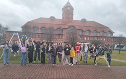 Na zdjęciu widać grupę uczniów pozujących przed budynkiem muzeum. Dzieci podskakują i uśmiechają się, co pokazuje ich radość i dobrą atmosferę podczas wycieczki. W tle znajduje się charakterystyczny ceglany budynek z wieżą oraz plac zabaw.