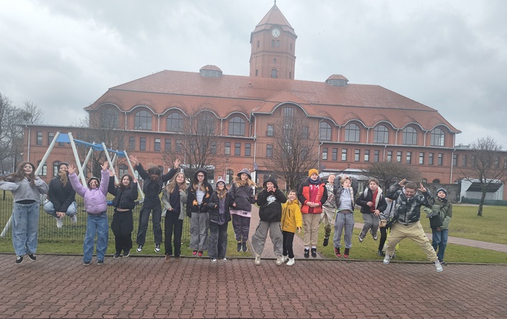 Na zdjęciu widać grupę uczniów pozujących przed budynkiem muzeum. Dzieci podskakują i uśmiechają się, co pokazuje ich radość i dobrą atmosferę podczas wycieczki. W tle znajduje się charakterystyczny ceglany budynek z wieżą oraz plac zabaw.