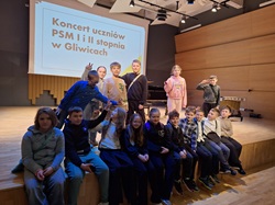 Na zdjęciu widać grupę uczniów klasy 4b, którzy pozują na scenie w sali koncertowej. Za nimi wyświetlony jest napis informujący o koncercie uczniów Państwowej Szkoły Muzycznej w Gliwicach. Dzieci siedzą i stoją razem, uśmiechają się i wyglądają na zadowolone z udziału w wydarzeniu.