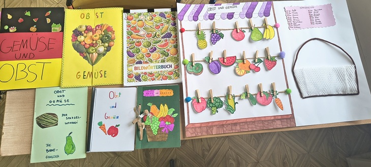 Na zdjęciu widać kolorowe prace uczniów przygotowane na konkurs – słowniczki obrazkowe dotyczące owoców i warzyw. Każda praca jest wykonana starannie i kreatywnie, z wykorzystaniem rysunków, wycinanek oraz różnych materiałów. Szczególnie wyróżnia się makieta z przypiętymi obrazkami owoców i warzyw, przypominająca mały stragan.