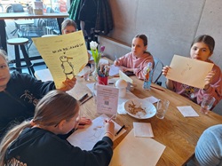 Na zdjęciu widać uczniów podczas warsztatów kulinarnych siedzących przy stole w restauracji. Dzieci rysują i prezentują swoje prace, a na stole znajdują się kredki, kartki oraz naczynia po posiłku. Atmosfera jest twórcza i swobodna, sprzyjająca zabawie i nauce