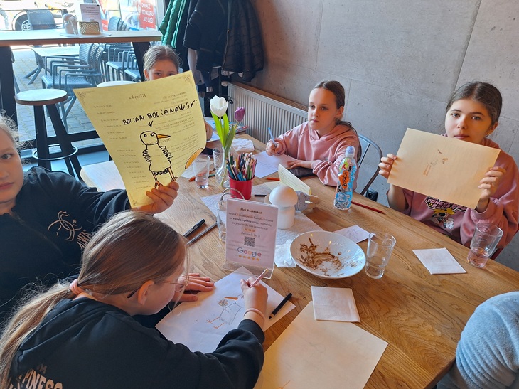 Na zdjęciu widać uczniów podczas warsztatów kulinarnych siedzących przy stole w restauracji. Dzieci rysują i prezentują swoje prace, a na stole znajdują się kredki, kartki oraz naczynia po posiłku. Atmosfera jest twórcza i swobodna, sprzyjająca zabawie i nauce