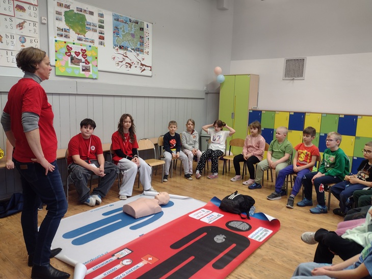 Na zdjęciu widać zajęcia szkolne poświęcone nauce udzielania pierwszej pomocy. W sali lekcyjnej grupa dzieci siedzi na krzesłach ustawionych w półkolu i uważnie słucha prowadzącej. Kobieta ubrana w czerwoną koszulkę stoi z boku i tłumaczy zasady postępowania w sytuacjach zagrożenia zdrowia lub życia.  Na podłodze rozłożone są specjalne maty szkoleniowe oraz fantom do ćwiczeń resuscytacji krążeniowo-oddechowej, co wskazuje, że uczniowie uczą się praktycznych umiejętności ratowniczych. Widać także elementy wyposażenia klasy, takie jak kolorowe szafki i plansze edukacyjne na ścianach.